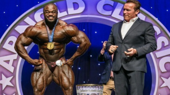 Brandon Curry smiles while posing with a gold medal on his bare chest while Arnold Schwarzenegger looks on in a suit and applauds.