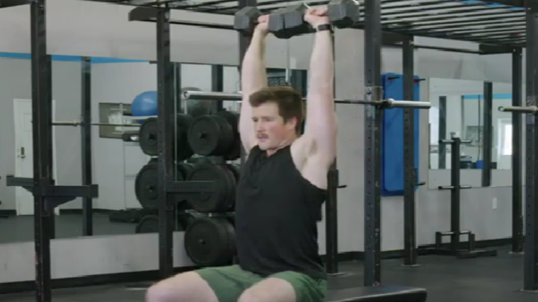 A person sits on a bench and presses two dumbbells overhead.