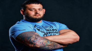 UK strongman Shane Flowers poses with his arms crossed in a blue WSM shirt against a black background.