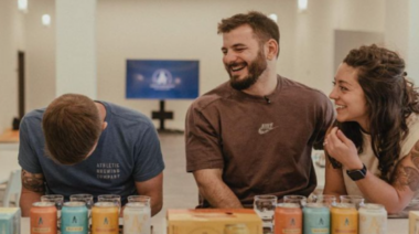 Mat Fraser sits with two people and laughs at a table filled with non-alcoholic beer cans.