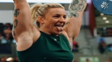 Kate Gordon sticks out her tongue and smiles while holding a loaded barbell overhead.