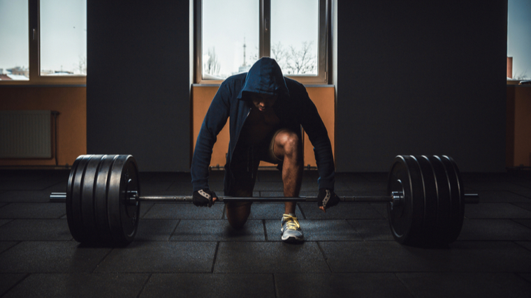 A person kneels on one knee in front of a barbell and looks down while wearing a blue hoodie.