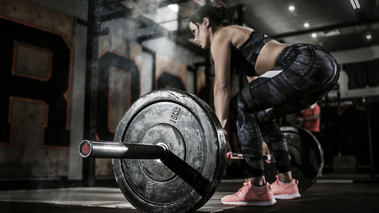 A person wears a sports bra, training pants, and pink shoes while preparing to lift a loaded barbell off the ground.