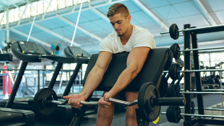 A person wearing a white t-shirt performs preacher curls in the gym.
