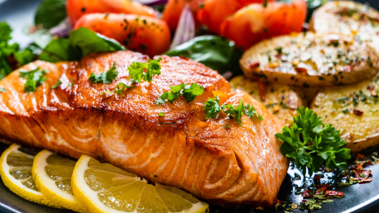 A full plate of food features cooked salmon, sliced potatoes, tomatoes, and a lemon slice.