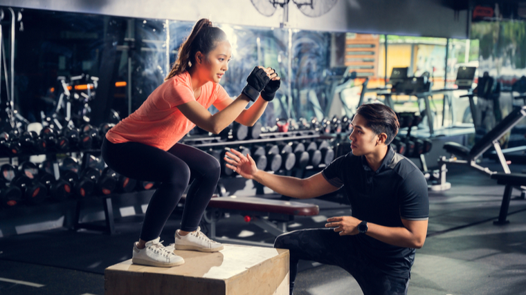 A person wearing a black shirt coaches a person wearing an orange shirt on how to perform a box jump.