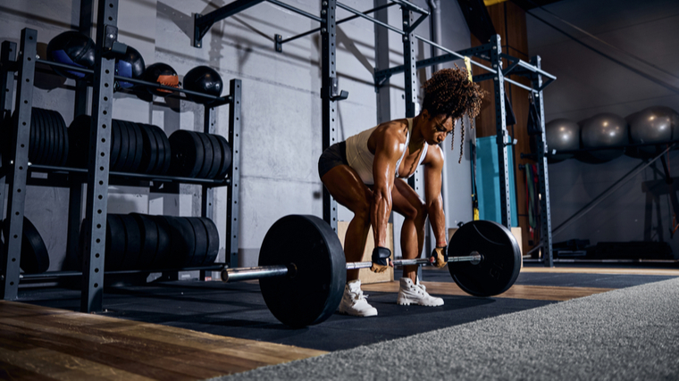 A person wears a white tank top and gym shorts while preparing to deadlift.