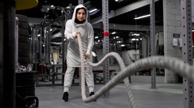 A person wears a beige long-sleeved hoodie and a hijab while doing battle rope waves.