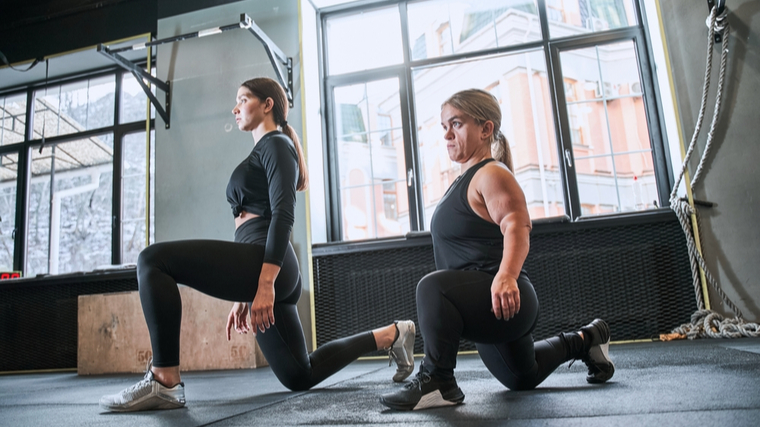 Two people wearing grey and black workout gear perform walking lunges in a gym.