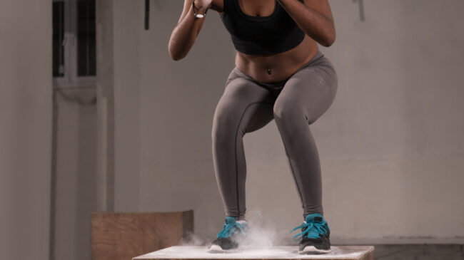 A person wearing a sports bra, grey leggings, and blue laces on their sneakers lands on a wooden plyo box as chalk spreads upward.