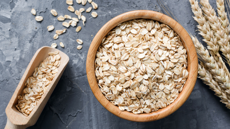 A wooden bowl of uncooked oats sits on a grey table alongside a wooden spoon spilling out.