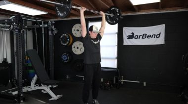Jake doing the standing barbell overhead press in the Barbend gym.