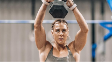Brooke Wells holds a dumbbell above her head with both hands.