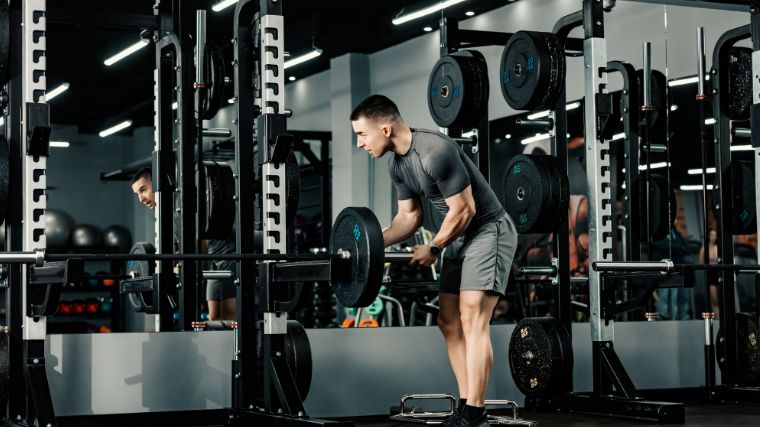 Man in grey t-shirt and shorts loads a weight plate onto a loaded barbell sitting on a power rack.