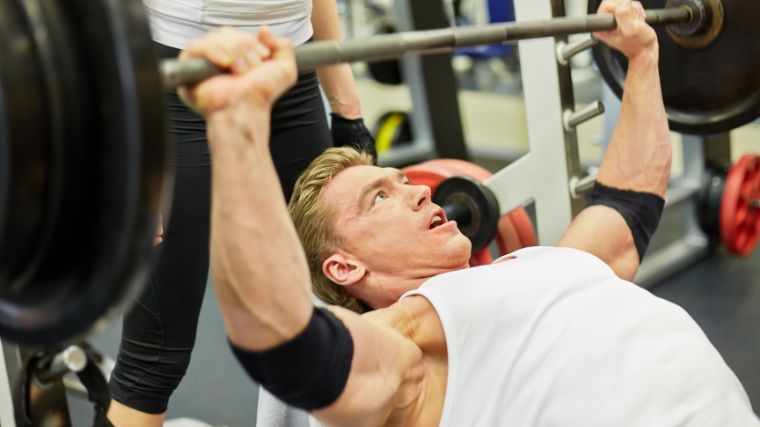 person wearing a white cut-off t-shirt and elbow sleeves incline bench presses a loaded barbell.
