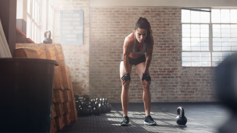 Person in blue sports bra and black shorts and lifting gloves bent over taking a rest in the gym