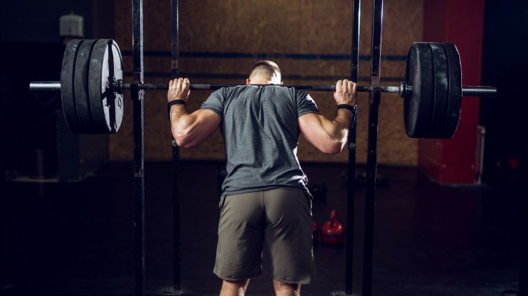 Person wearing a blue t-shirt and grey shorts placing their traps under a loaded barbell set in a power rack