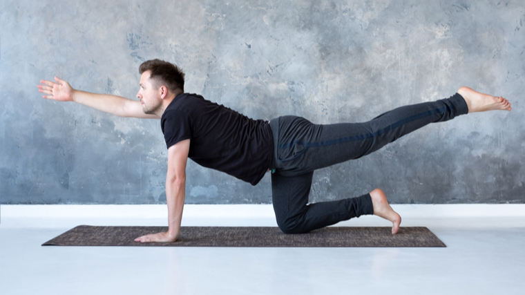 A person wearing a black t-shirt and grey yoga pants performs a bird dog.