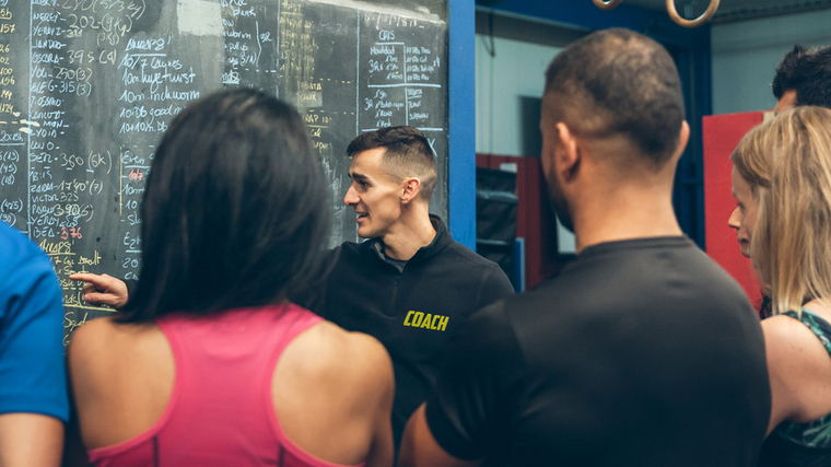 A person wearing a black polo that says "coach" in yellow print points to a chalkboard while talking to clients in a gym.