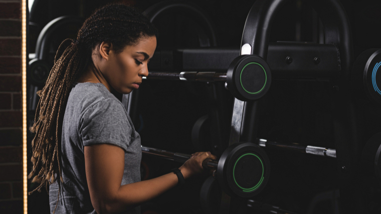 A person with braids wears a grey t-shirt and selects a barbell from a weight rack.