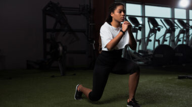 A person wearing loose braids, a white t-shirt, and black leggings performs a bodyweight lunge in the gym.