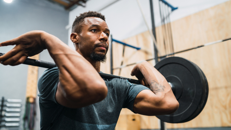 A person wears a blue-grey t-shirt and holds a loaded barbell in a front rack position.