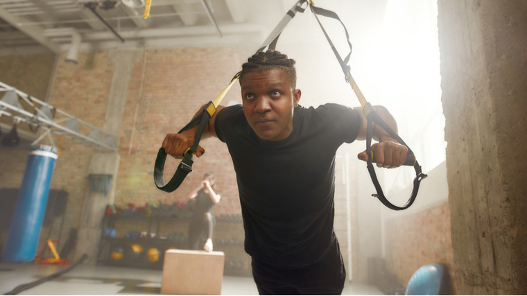 A person with a fade and cornrows wears a black t-shirt while training with a TRX suspension.
