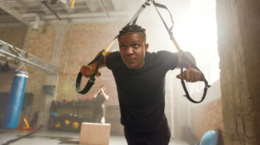 A person with a fade and cornrows wears a black t-shirt while training with a TRX suspension.