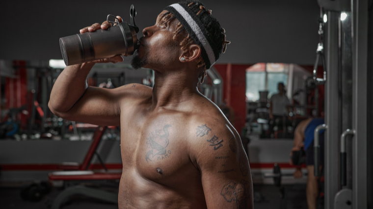 A shirtless person wearing a headband drinks from a shaker bottle in the gym.