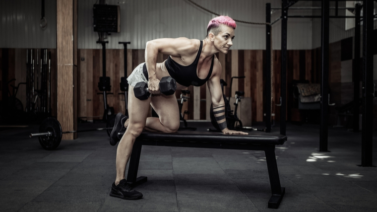 A person with short pink hair wears a sports bra while performing a single-arm dumbbell row.