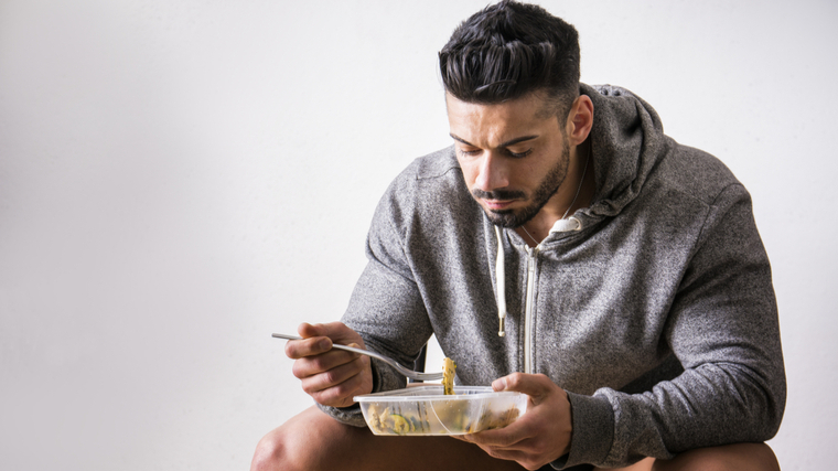 A person with facial hair wears a gray hoodie and shorts while eating food from a tupperware.