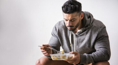 A person with facial hair wears a gray hoodie and shorts while eating food from a tupperware.