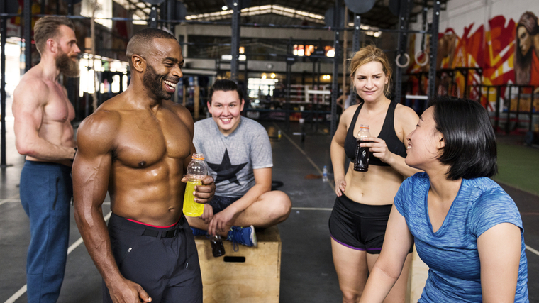 Several people sit and stand in a gym, talking and laughing after a workout.