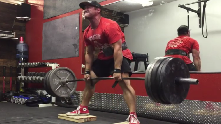 A person wearing a red Westside Barbell shirt, lifting belt, and baseball cap performs a deficit sumo deadlift.