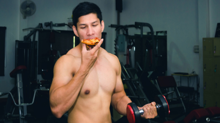 A muscular shirtless person takes a bite of pizza while holding a dumbbell in the gym.