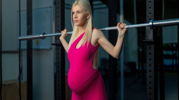 A pregnant person performs a back squat while wearing a pink exercise jumpsuit.
