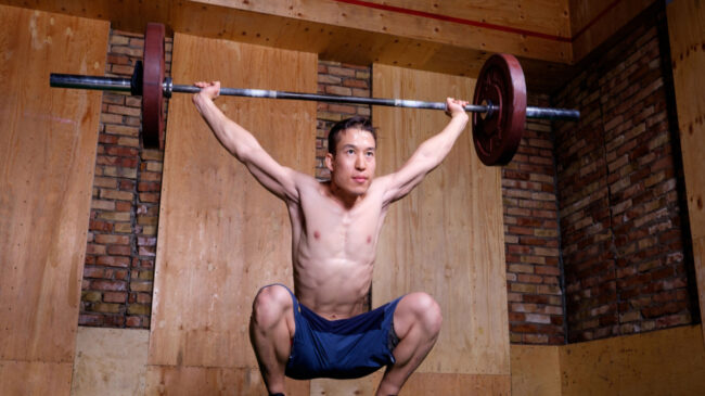 A shirtless person performs an overhead squat in a wood- and brick-walled gym.