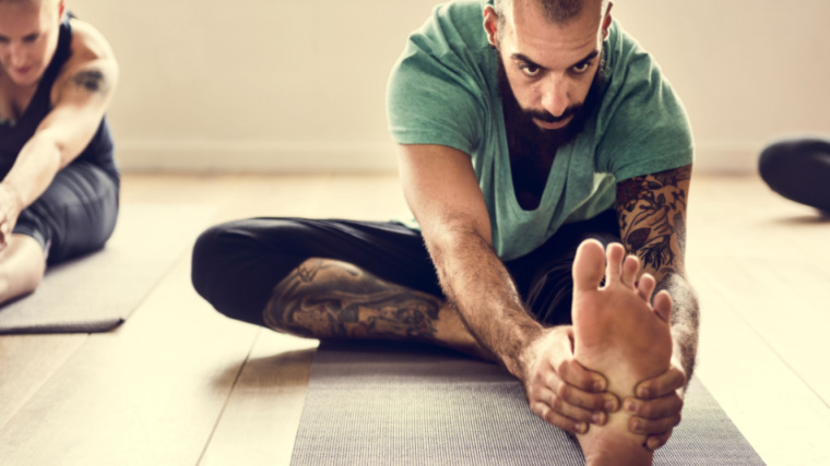 A person with tattoos stretches alongside a fellow yoga classmate.