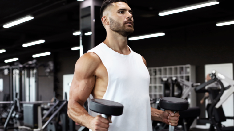 A person with a fresh haircut and facial hair wears a white cut-off sleeveless shirt while performing hammer curls with two dumbbells.