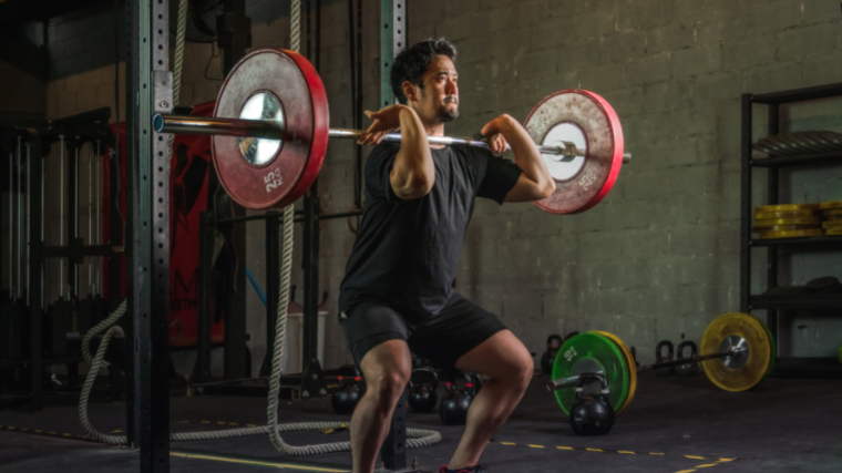A person wearing a black t-shirt and black shorts grimaces slightly as they perform a front squat with a loaded barbell.