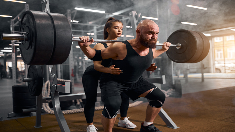 A person wears a tank top and knee sleeves while squatting heavy and receiving a spot from someone behind him.