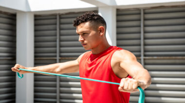 A person wearing a red tank top performs band pull-aparts at an outdoor gym.