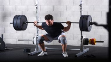 A person wears a black t-shirt and grey knee sleeves while squatting a loaded barbell.