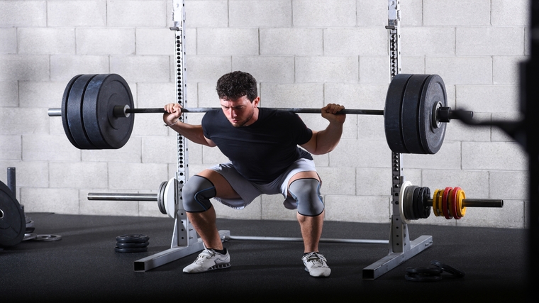 A person wears a black t-shirt and grey knee sleeves while squatting a loaded barbell.