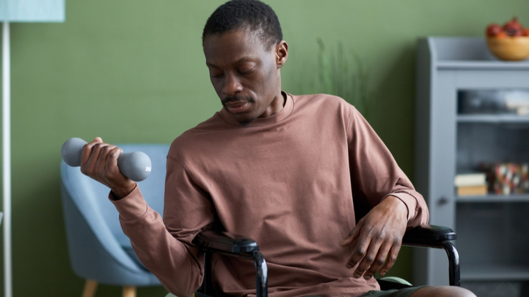 A person performs a dumbbell curl while sitting in a wheelchair at home.