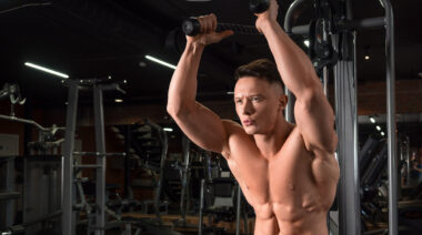 A shirtless bodybuilder performing overhead cable triceps extensions.