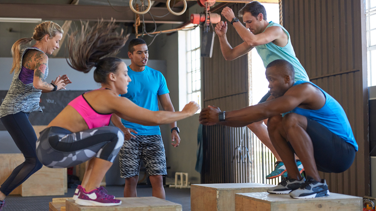 Four people perform box jumps while a fifth person stands by and coaches them.