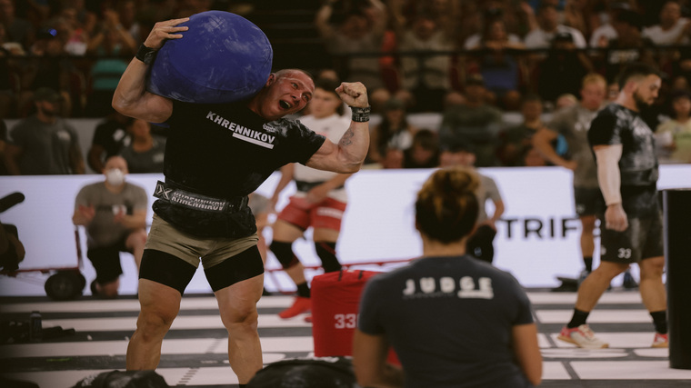 Roman Khrennikov celebrates a successful lift by flexing one arm while the other holds a sandbag on his shoulder.