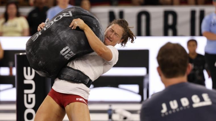 Tia-Clair Toomey, wearing red shorts and a white shirt, hugs a heavy sand bag tightly to her chest.