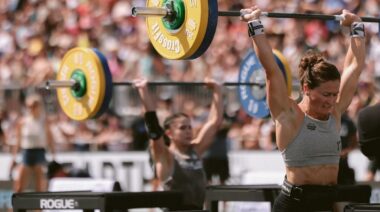Two women jerking barbells overhead, wearing tank tops and shorts.